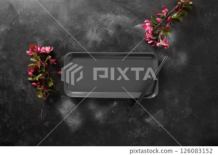 Minimalist scene of empty rectangular plate for asian food with black chopsticks on dark textured surface with spring pink flowers. Asian style. View from above. Copy space. Japanese food concept. Minimalist scene of empty rectangular plate for asian food with black chopsticks on dark textured surface with spring pink flowers. Asian style. View from above. Copy space. Japanese food concept. 126083152