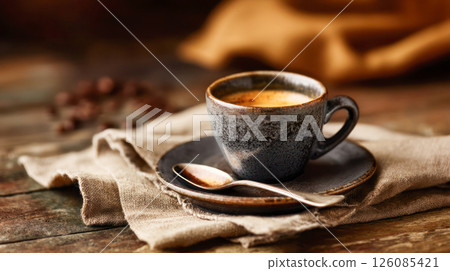 Coffee cup filled with rich espresso on a rustic wooden table, accompanied by a silver spoon and scattered coffee beans, creating a cozy atmosphere 126085421