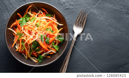 Fresh vegetable salad with vibrant colors and textures, featuring shredded carrots, cabbage, and herbs, served in a dark bowl on a textured surface 126085423