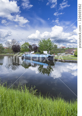 Houseboat reflecting on the calm water of a canal in the French countryside under a blue sky with white clouds Houseboat reflecting on the calm water of a canal in the French countryside under a blue sky with white clouds 126085982