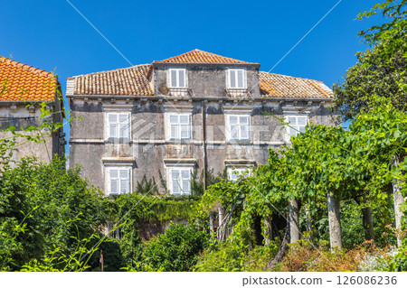 Orebic town at coast of the Peljesac peninsula in Croatia, Europe. Historic European villa with shuttered windows and tiled roof, surrounded by lush vegetation and a clear blue sky. 126086236