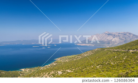Sea coast of the Peljesac Peninsula in Croatia, Europe. Coastal vista featuring a striking mountain range, deep blue waters, lush green hills, and a serene, clear blue sky on a sunny day. 126086304