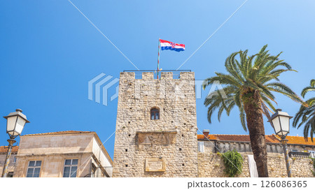 Korcula town in island of Korcula in Adriatic sea in Croatia, Europe. A historical stone tower stands tall against a clear blue sky, topped with a national flag and framed by palm trees. Korcula town in island of Korcula in Adriatic sea in Croatia, Europe. A historical stone tower stands tall against a clear blue sky, topped with a national flag and framed by palm trees. 126086365