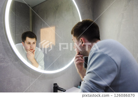 Man in a light sweater leans over a bathroom sink, thoughtfully examining his reflection in a round LED-lit mirror during his morning routine. 126086870