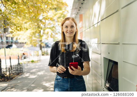 Young Woman Using Parcel Locker with Smartphone Outdoors 126088100