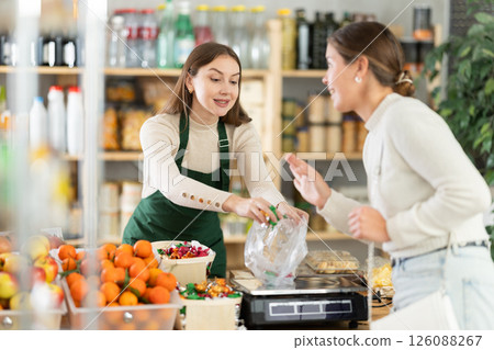 Woman sells and puts candies in bag for young woman, puts them on scale and weighs fruits Woman sells and puts candies in bag for young woman, puts them on scale and weighs fruits 126088267