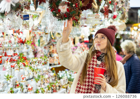 Girl with glass of coffee to go looks at wreaths and garlands of pine cones and red berries 126088736