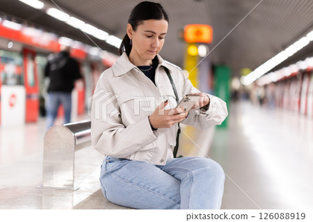 Woman monitors navigation of train in smartphone sitting on a bench at metro station Woman monitors navigation of train in smartphone sitting on a bench at metro station 126088919