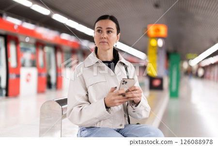 Woman monitors navigation of train in smartphone sitting on a bench at metro station 126088978