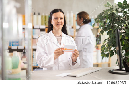 Middle-aged female pharmacist offering pills box while man working in drugstore 126089085