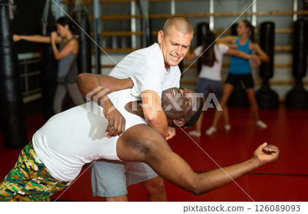 Caucasian and African-american men sparring to practice neck grabbing in gym during self-protection training. Women exercising in background. 126089093