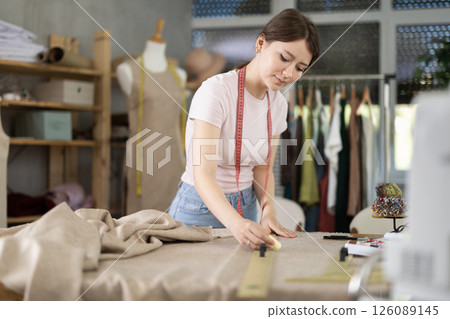 Girl tailor works in sewing workshop - draws lines on fabric using chalk according to pattern 126089145