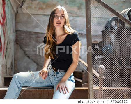 Woman in jeans and black tee shirt posing near steel mesh fencing in destroyed plant 126089172