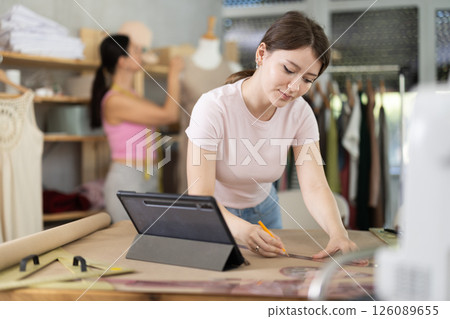 Girl tailor draws dress pattern of product on paper, tablet on table. Companion in background Girl tailor draws dress pattern of product on paper, tablet on table. Companion in background 126089655