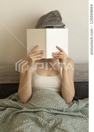 woman with a book sitting in bed with a towel on her head 126089664