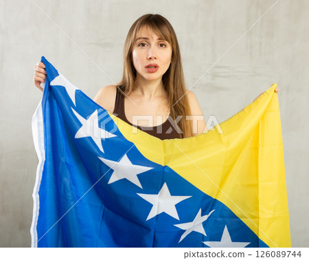 Sad young woman holding flag of Bosnia and Herzegovina against unicoloured background Sad young woman holding flag of Bosnia and Herzegovina against unicoloured background 126089744