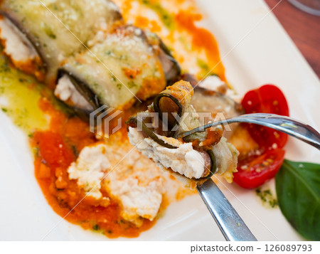 Eggplant rolls baked in gravy stuffed with soft cheeses lie on plate with pieces of fresh vegetables Eggplant rolls baked in gravy stuffed with soft cheeses lie on plate with pieces of fresh vegetables 126089793