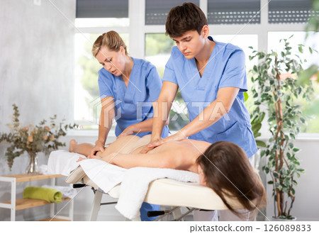 Female professional physiotherapist helps guy student to perform back massage to female client. Female professional physiotherapist helps guy student to perform back massage to female client. 126089833