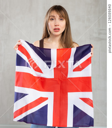 Upset patriotic young female student holding state flag of Great Britain against gray wall background indoors 126089840