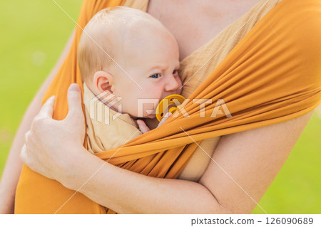 Mother holding her little baby in a yellow sling in the park. Warm and loving family moment. Babywearing, parenting, and mother-child bonding concept Mother holding her little baby in a yellow sling in the park. Warm and loving family moment. Babywearing, parenting, and mother-child bonding concept 126090689