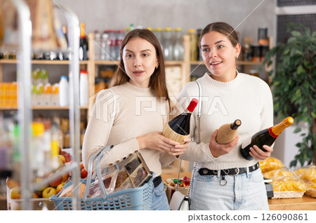 Two girls in store while shopping, choose bottle of wine and chat sweetly Two girls in store while shopping, choose bottle of wine and chat sweetly 126090861