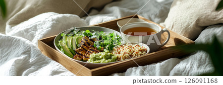 A wholesome breakfast scene with quinoa, grilled tofu, avocado, and microgreens, served with mint tea on a dark wooden tray, set on a white linen bed in a cozy, light-filled bedroom, banner 126091168