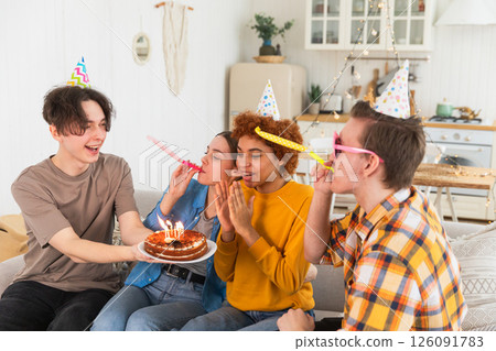 Make a wish. Woman wearing party cap blowing out burning candles on birthday cake. Happy Birthday party. Group of friends wishes girl happy birthday. People celebrating birthday with party at home 126091783
