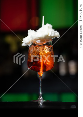 A vibrant orange cocktail, likely a spritz, is served with ice in a tall glass and topped with a fluffy white cloud of cotton candy and a white straw 126091970