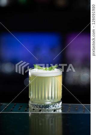 A pale yellow cocktail with a frothy white top is garnished with two vibrant green mint leaves, sitting on a reflective bar surface. 126091980