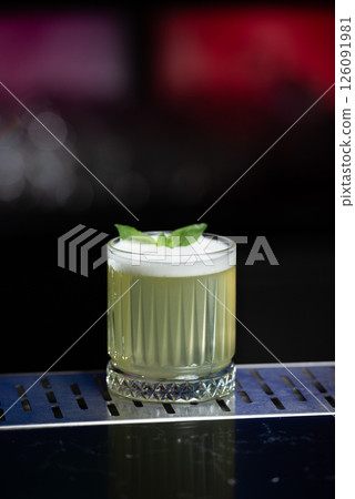 A pale yellow cocktail with a frothy white top is garnished with two vibrant green mint leaves, sitting on a reflective bar surface. 126091981