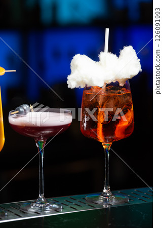 Two vibrant cocktails sit on a bar counter, one a refreshing green mix with mint and a straw, the other a bright orange topped with white sugar cotton candy and a straw. 126091993