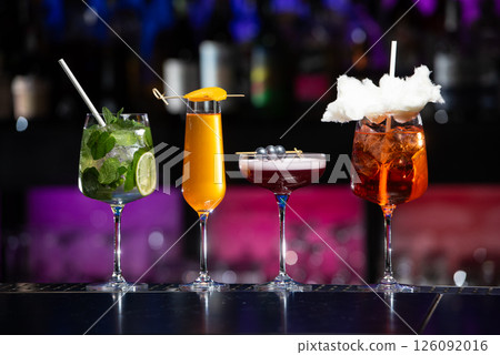 Five distinct and colorful cocktails are lined up on a sleek bar counter, each uniquely garnished with ingredients like mint, citrus slices, blueberries, and cotton candy 126092016