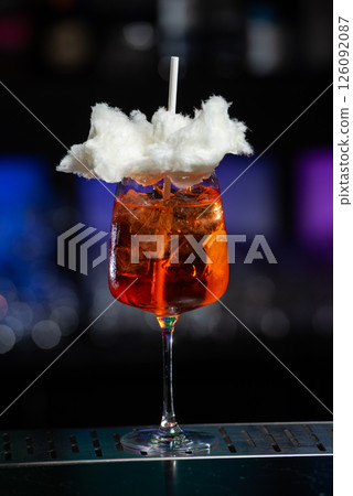 A vibrant orange cocktail filled with ice sits on a textured bar, topped with a whimsical cloud of white cotton candy and a white straw. 126092087