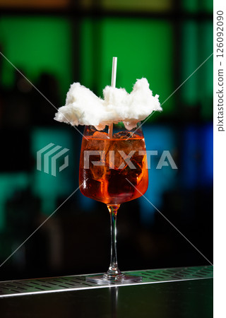 A vibrant orange cocktail filled with ice sits on a textured bar, topped with a whimsical cloud of white cotton candy and a white straw. A vibrant orange cocktail filled with ice sits on a textured bar, topped with a whimsical cloud of white cotton candy and a white straw. 126092090