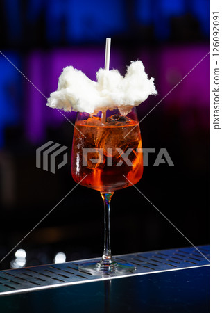 A vibrant orange cocktail filled with ice sits on a textured bar, topped with a whimsical cloud of white cotton candy and a white straw. A vibrant orange cocktail filled with ice sits on a textured bar, topped with a whimsical cloud of white cotton candy and a white straw. 126092091