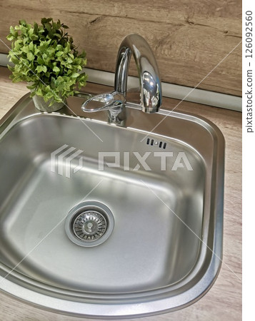 Stainless steel kitchen sink with modern faucet and green plant in wooden interior. Bright and clean eco-style home detail Stainless steel kitchen sink with modern faucet and green plant in wooden interior. Bright and clean eco-style home detail 126092560