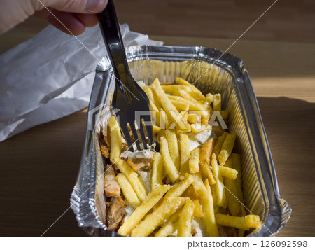 Person holding takeout container with kebab, french fries, drizzling white garlic sauce and using black plastic fork for eating 126092598