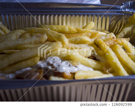 Street food delicacy featuring crispy fries layered over juicy kebab meat, smothered in creamy garlic sauce, served in disposable aluminum container for convenient dining Street food delicacy featuring crispy fries layered over juicy kebab meat, smothered in creamy garlic sauce, served in disposable aluminum container for convenient dining 126092599
