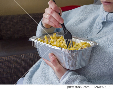 Cozy woman relaxing in pajamas, munching kebab and fries from takeout container while lounging on living room sofa 126092602