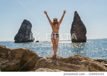Woman Beach Rocks - A woman in a bikini stands on a rock with her arms raised, facing the ocean and two large rocks in the distance. 126093281