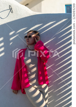 Here's a title attempting to follow all instructions: Cardigan Fashion Greece Summer: Woman poses in pink cardigan against whitewashed wall, showcasing summer style in Greece. 126093289