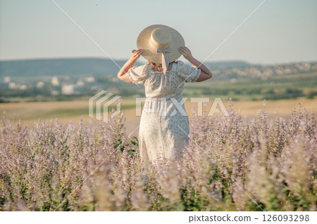 Woman Hat Field: Summer rural landscape; woman in white dress carries hat, walking through purple wildflowers for sun protection. Woman Hat Field: Summer rural landscape; woman in white dress carries hat, walking through purple wildflowers for sun protection. 126093298