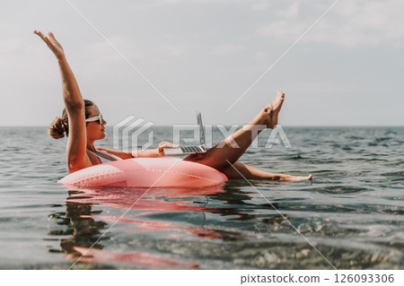 A woman is floating on a pink inflatable raft in the ocean while using a laptop. Concept of relaxation and leisure, as the woman enjoys her time in the water while working on her laptop. 126093306