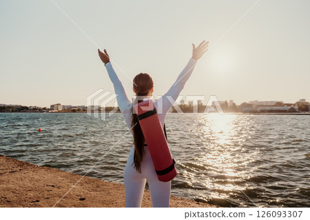 Yoga Woman Beach Sunset - A woman in white clothing stands on a beach with her arms raised high in the air, looking at the setting sun over the water while holding a pink yoga mat. 126093307