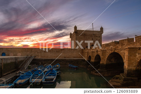 Blue fishing boats in a harbor with foretress bridge and Port of Essaouira Sqala, Portuguese castle in a sunset lights, Essaouira, Morocco 126093889