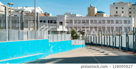 Urban outdoor path with modern fences and buildings under a bright blue sky. Urban outdoor path with modern fences and buildings under a bright blue sky. 126095627