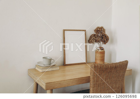 Minimalist home workspace with wooden desk, ceramic cup of tea, coffee styled in warm earthy tones. Dried flowers in vase. Empty picture frame, poster mockup. Framed print. Cozy interior inspiration 126095698