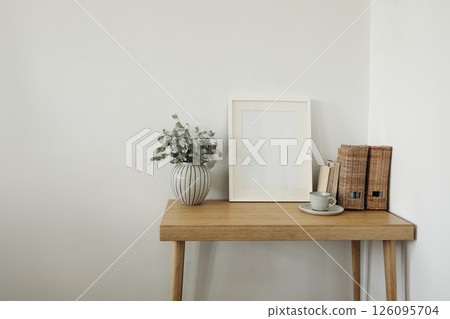 Elegant Scandinavian interior. Green dry eucalyptus tree branches in vase on desk, table. Cup of coffee tea, old books. Empty white vertical picture frame mockup, wall background. Wicker magazine file 126095704