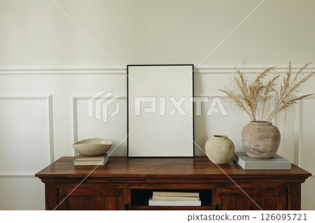 Elegant living room. Mahogany wooden cabinet dresser. Empty picture mock up poster frame. Clay vases, dry grass bouquet, ceramic bowl. Modern Scandinavian interior design, white wall background. 126095721