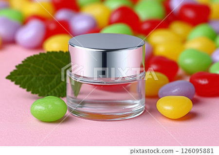 Small, clear cosmetic bottle with a silver cap sits on a pink surface, surrounded by colorful candies and accompanied by a vibrant green leaf, creating a playful and appealing presentation 126095881